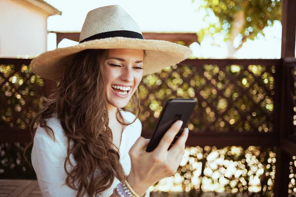 在宾馆的阳台上 身穿白衬衫 头戴帽子 头戴智能手机 面带微笑的40多岁的家庭主妇正在用智能手机与客人见面 — 图库照片