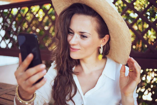 ホワイトシャツを着た幸せな現代女性がゲストハウスホテルのテラスでスマートフォンでオンライン会議をしていて — ストック写真