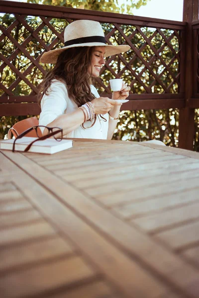 穿着白衬衫 头戴咖啡杯 帽子和眼镜的现代中年家庭主妇坐在阳台桌旁笑着 — 图库照片