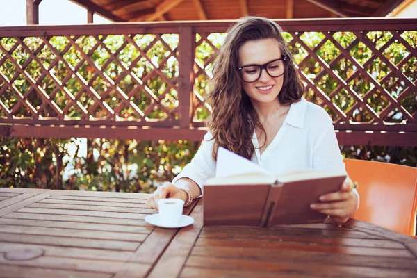在宾馆的阳台上 身穿白衬衫 头戴眼镜 面带微笑的优雅女人坐在餐桌边喝咖啡看书 — 图库照片