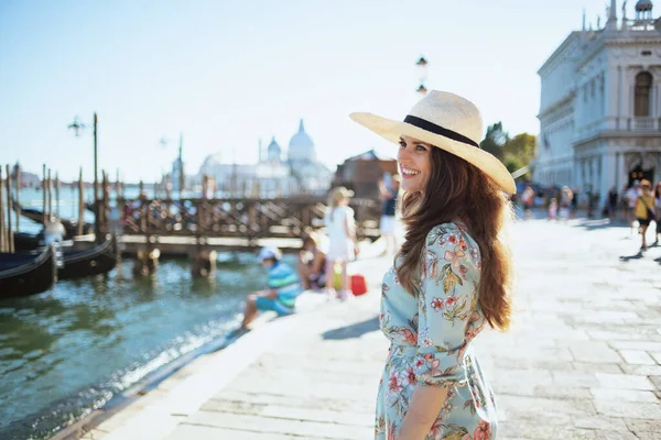 イタリアのヴェネツィアで堤防の遠足を持っている帽子と花のドレスで流行のソロ観光客の女性を笑顔 — ストック写真