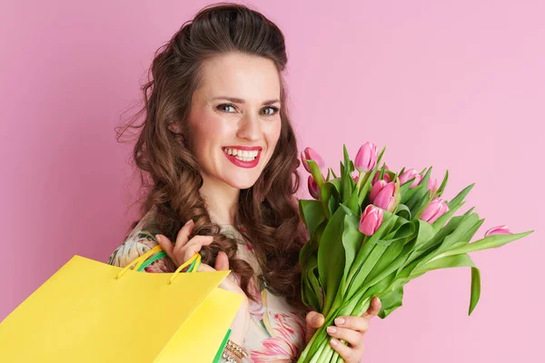 笑顔スタイリッシュな中年女性とともに長い波状ブルネット髪でチューリップ花束とショッピングバッグ隔離されたピンク — ストック写真