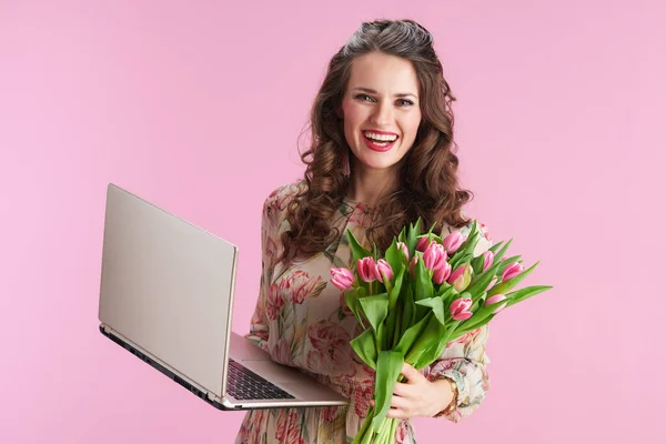 笑顔現代的な中年女性で花のドレスでチューリップ花束とラップトップ隔離されたピンクの背景 — ストック写真