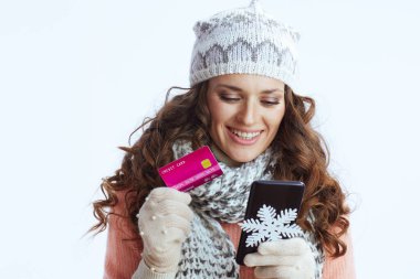 Merhaba kış. Süveter, eldiven, şapka ve eşarp giymiş gülümseyen modern bir kadın kar tanesi, akıllı telefon ve kredi kartı ile beyaz arka planda e-ticaret sitesinde online alışveriş yapıyor..