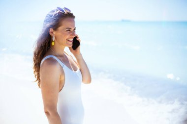 Mutlu modern 40 yaşında plajda beyaz plaj elbiseli bir kadın akıllı telefondan konuşuyor..