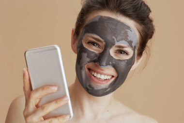 Kozmetik maskeli mutlu orta yaşlı kadın bej arka planda izole akıllı telefon kullanıyor..