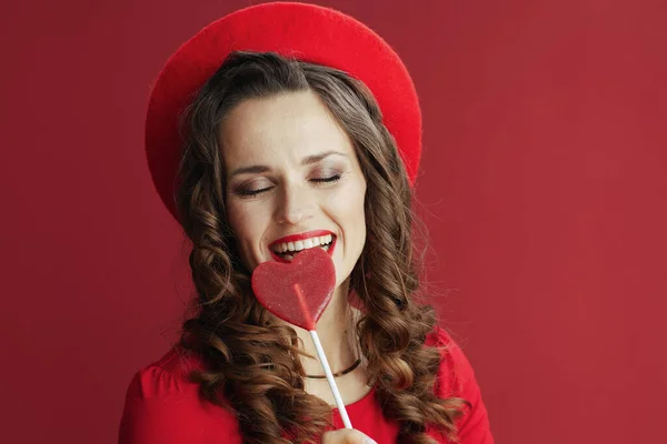 ハッピーバレンタイン 幸せなスタイリッシュな中年女性で赤ドレスとベレー帽隔離上の赤の背景にハート形のキャンディーオンスティック — ストック写真
