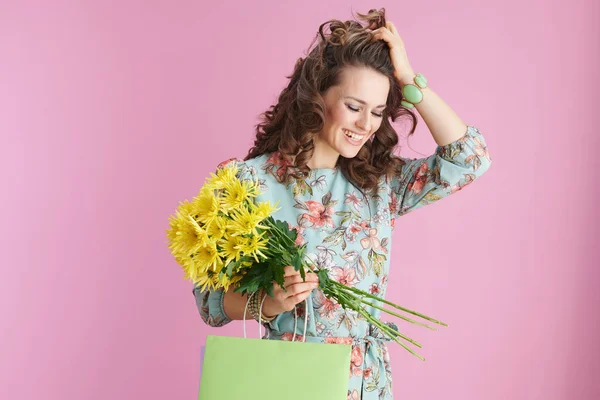 幸せな中年齢女性とともに長い波状ブルネット髪とともに黄色菊の花と緑のショッピングバッグ隔離されたピンク — ストック写真