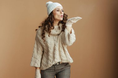 Merhaba kış. Bej kazaklı, eldivenli ve şapkalı zarif bir kadın bej arka planda kar üflüyor..