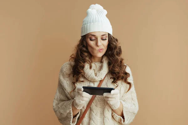 Hello winter. pensive trendy woman in beige sweater, mittens and hat on beige background using smartphone applications.