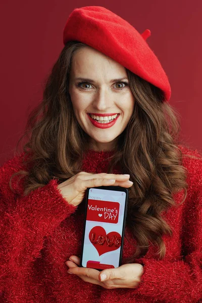 Happy Valentine. happy elegant middle aged woman with long wavy hair in red sweater and beret , smartphone and Valentine app.