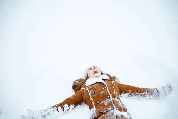 happy modern 40 years old woman in brown hat and scarf in sheepskin coat with mittens making snow angels outside in the city in winter.