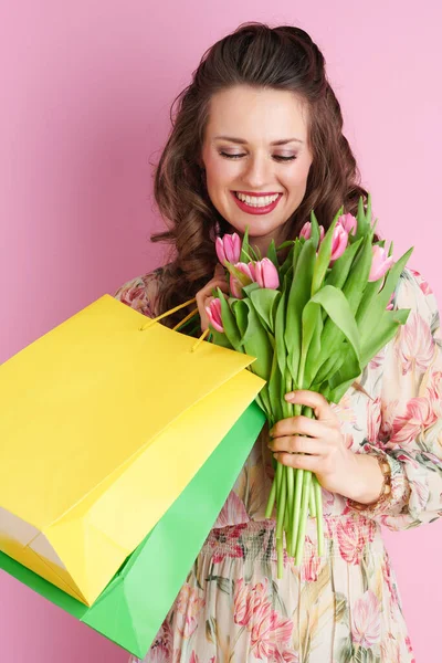 幸せな若いです若いです女性とともに長い波状ブルネット髪でチューリップ花束とショッピングバッグ隔離されたピンクの背景 — ストック写真