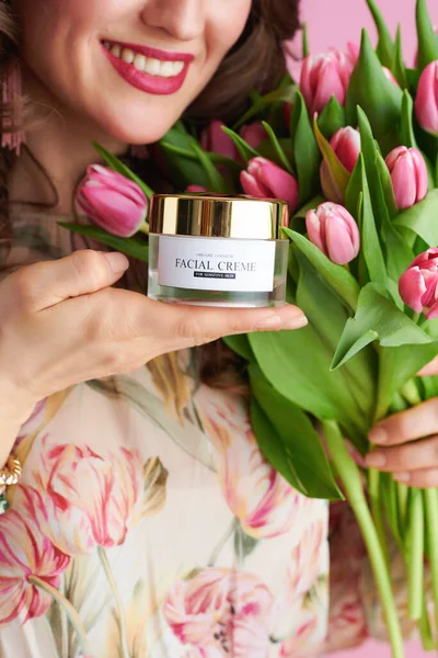 Closeup on smiling woman with tulips bouquet and cosmetic jar isolated on pink.