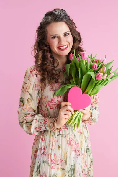 ピンク色に孤立したチューリップの花束とピンクのハートを持つ花のドレスの現代女性の笑顔 — ストック写真