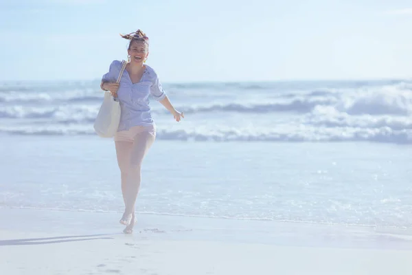 Full length portrait of happy elegant female with white straw bag walking at the beach.
