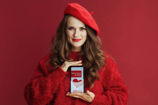 Happy Valentine. happy elegant 40 years old woman with long wavy hair in red sweater and beret , smartphone and Valentine app.