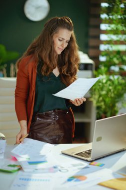 smiling young small business owner woman with laptop working with documents in the modern green office.