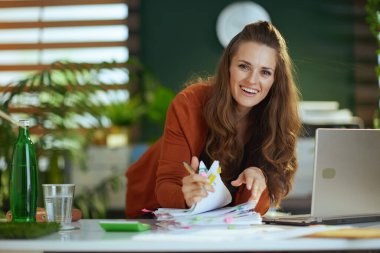happy stylish middle aged small business owner woman with laptop working with documents in the modern green office.