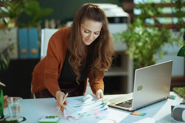 happy stylish 40 years old small business owner woman with laptop working with documents in the modern green office.
