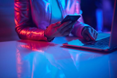 Neon metaverse futuristic concept. Closeup on female with laptop using smartphone applications in office.