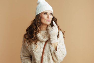 Hello winter. modern 40 years old woman in beige sweater, mittens and hat with hygienic lipstick isolated on beige.