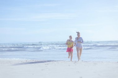 Sahilde koşan gülümseyen modern anne ve genç kızın tam boy portresi..