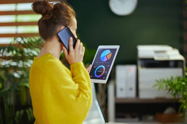 Seen from behind small business owner woman in yellow sweater in the green office speaking on a smartphone, using tablet PC and studying charts.