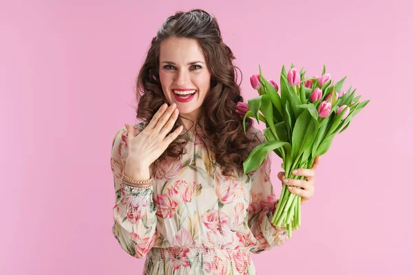 confused stylish 40 years old woman with long wavy brunette hair with tulips bouquet against pink background.