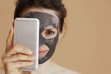 modern 40 years old woman with cosmetic mask using smartphone isolated on beige background.