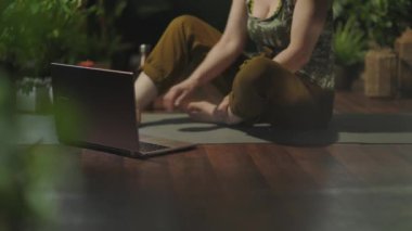 Yeşil evde dizüstü bilgisayarlı bir kadının yoga yapmasına yakın çekim..