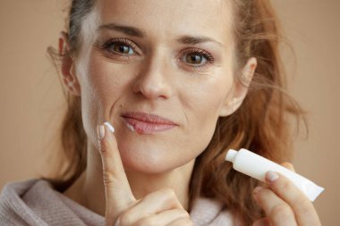 40 years old woman with herpes on lips and ointment in a tube applying ointment using finger against beige background.