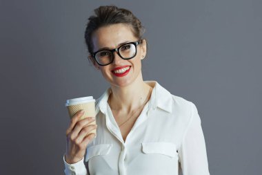 happy trendy 40 years old woman employee in white blouse with glasses and coffee cup isolated on gray background.