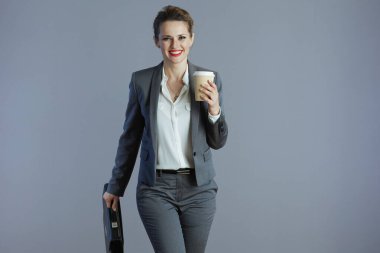 smiling stylish small business owner woman in gray suit with coffee cup and briefcase walking against grey background.