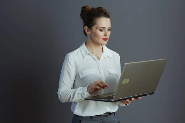 modern 40 years old woman employee in white blouse using laptop on grey.