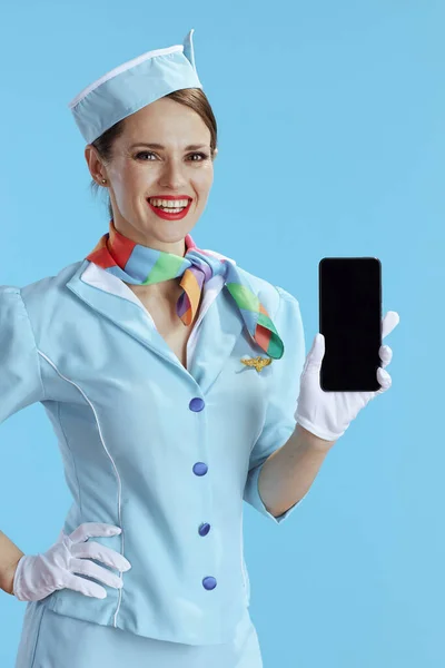 happy elegant female flight attendant on blue background in blue uniform showing smartphone blank screen.