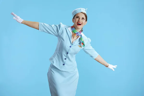 happy elegant female air hostess against blue background in blue uniform with outstretched arms imitating flight of airplane.
