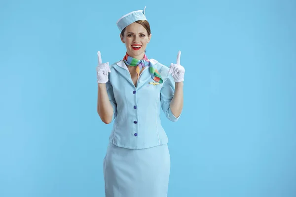 smiling stylish female air hostess on blue background in blue uniform pointing up at copy space.
