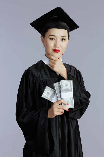 smiling modern female asian graduate student with money isolated on gray.
