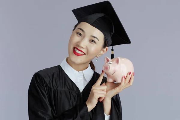 smiling modern female asian graduate student with piggy bank against gray background.
