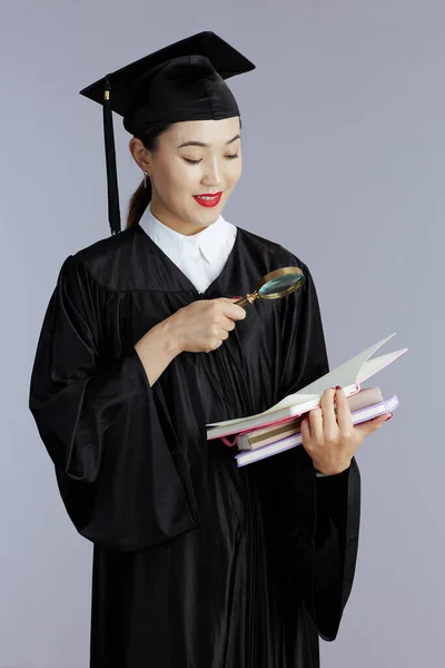 happy young female asian graduate student with books, notebooks and magnifying glass isolated on gray background.