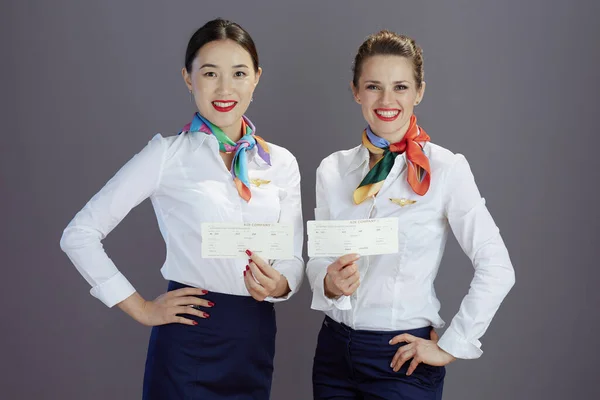 happy modern female flight attendants in blue skirt, white shirt and scarf with flight tickets against grey background.
