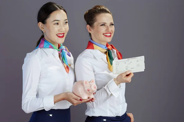 happy stylish flight attendant women in blue skirt, white shirt and scarf with flight tickets and piggy bank isolated on gray background.