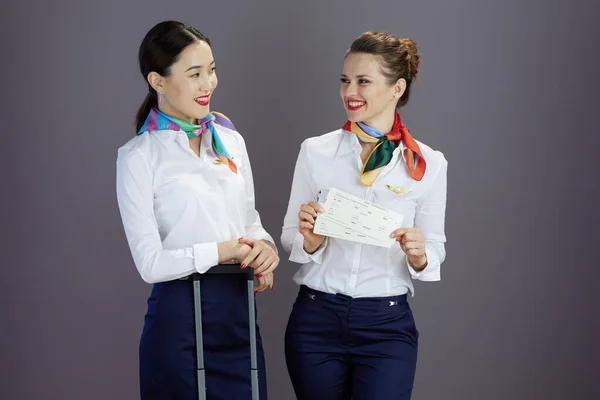 happy elegant female air hostesses in blue skirt, white shirt and scarf with flight tickets and travel bag against gray background.
