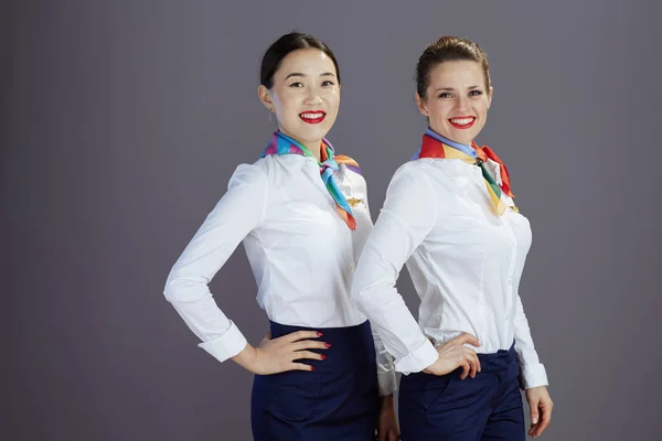 Portrait of happy modern air hostess women in blue skirt, white shirt and scarf against grey background.