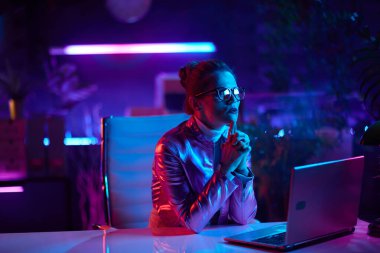 Neon metaevren fütüristik konsepti. Modern ofiste dizüstü bilgisayarlı gözlüklü şık orta yaşlı kadın..