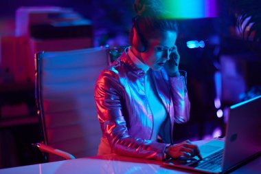 Neon metaevren fütüristik konsepti. Modern ofiste dizüstü bilgisayarı ve kulaklığı olan şık orta yaşlı bir kadın..