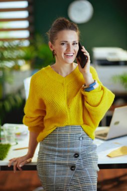 Gülümseyen modaya uygun küçük işletme sahibi sarı kazaklı kadın modern yeşil ofiste akıllı bir telefonla konuşuyor..