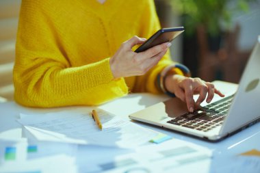 Closeup on small business owner woman in yellow sweater with laptop using smartphone in the green office. clipart