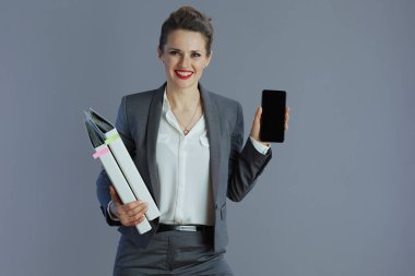 Gülümseyen orta yaşlı, gri takım elbiseli, gri arkaplanda izole akıllı telefon boş ekranı gösteren bir iş kadını..